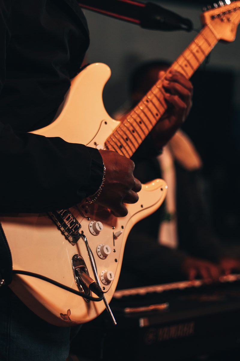 Un guitariste joue sur une guitare blanche, un autre musicien en arrière-plan.